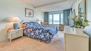 Une chambre à coucher comprenant un lit, des tables de chevet, une commode, une chaise, une fenêtre donnant sur la plage et une plante.