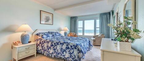 A bedroom with a bed, bedside tables, a dresser, a chair, a window with a view of the beach, and a plant.