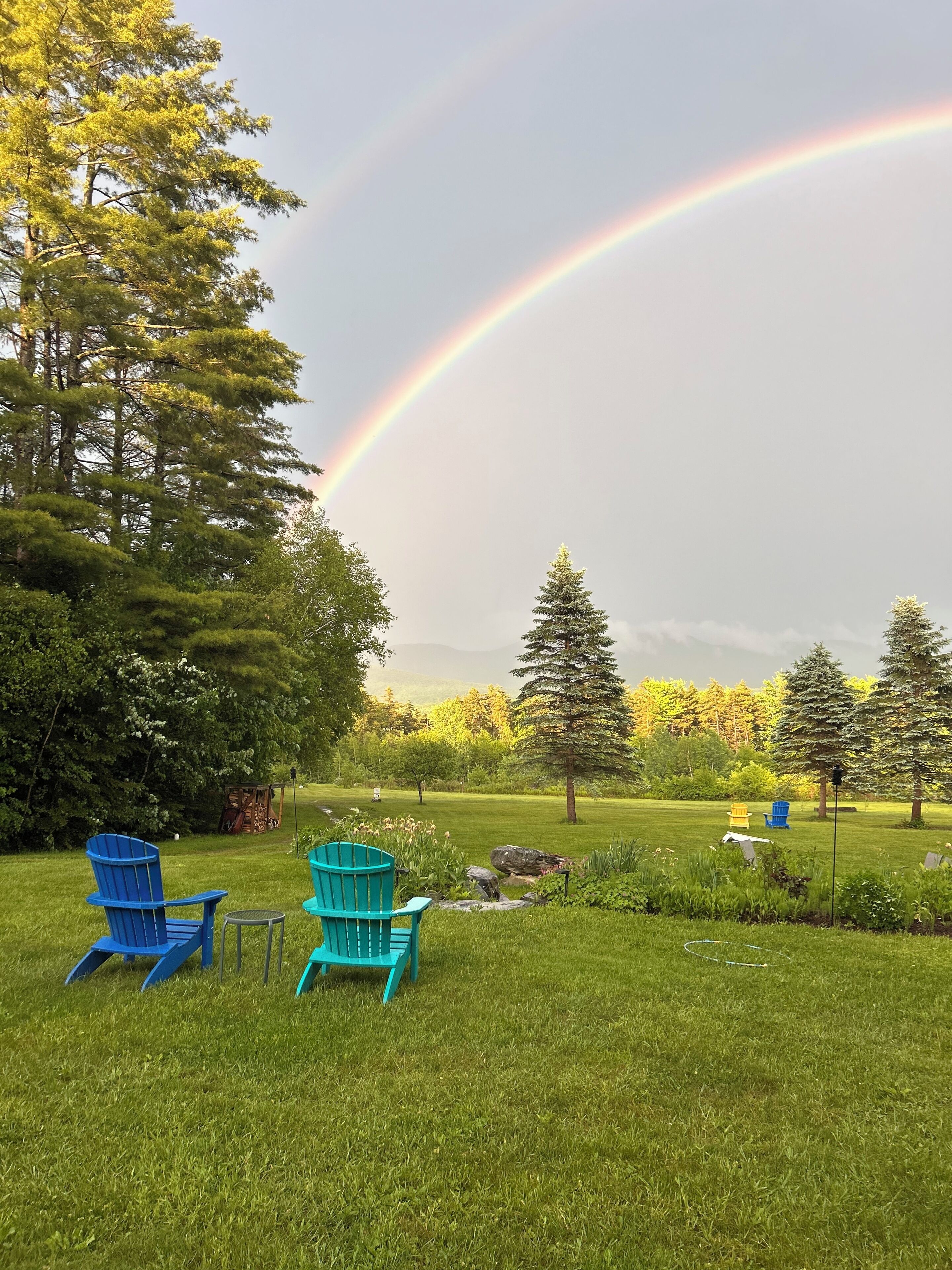 A grassy outdoor area with two blue chairs and a small table, surrounded by trees and a visible rainbow.