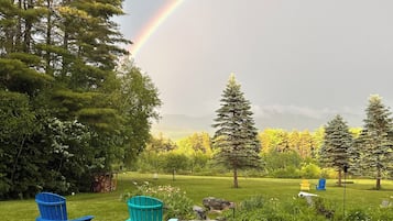 A grassy outdoor area with two blue chairs and a small table, surrounded by trees and a visible rainbow.