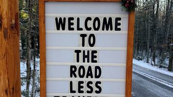 A welcome sign with the text 'WELCOME TO THE ROAD LESS TRAVELED' mounted on a wooden post.