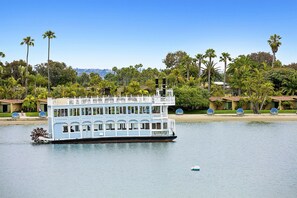 A large, multi-deck houseboat with a white exterior and blue accents, docked near a shoreline with palm trees and houses. - Bay Front Luxury Penthouse w/elev. & AC! Enjoy multiple outside patios & views! (San Diego)