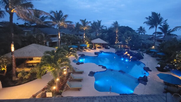 A resort pool area with lounge chairs and umbrellas, surrounded by palm trees and a thatched-roof building. - Hawaiian Escape with all the Amenities  (Kapolei)