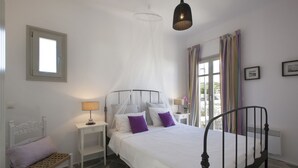 Une chambre à coucher comprenant un lit, des tables de chevet, un banc, une fenêtre avec des rideaux et un luminaire suspendu.