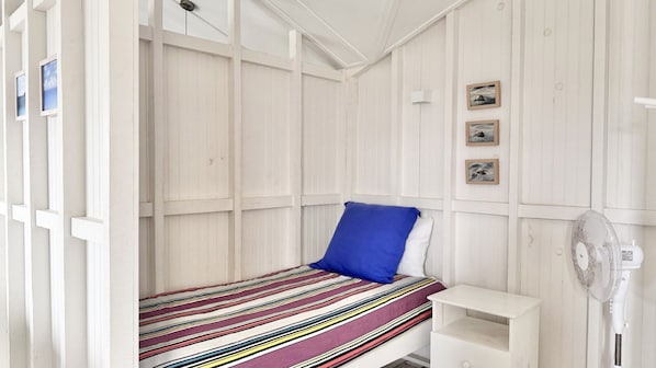 Ein Zimmer mit einem Einzelbett, einer gestreiften Tagesdecke, einem blauen Kopfkissen und einem weißen Nachttisch. An der Wand hängen drei gerahmte Bilder.