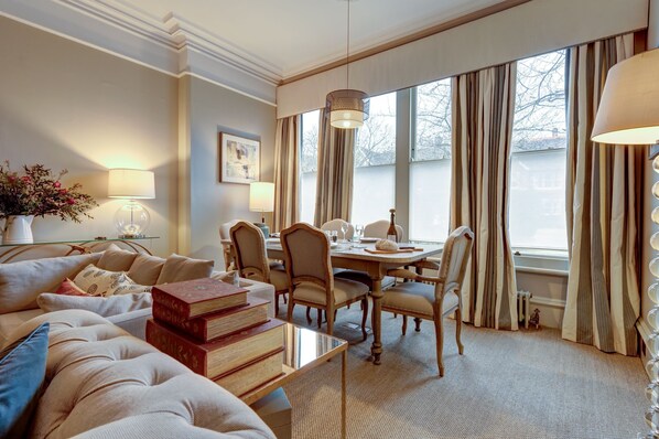 Ein stilvoll eingerichtetes Hotelzimmer mit einer gemütlichen Sitzecke, einem Esstisch und großen Fenstern mit Vorhängen.