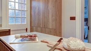 A room with a wooden door, a window with a grid pattern, a table with a book, a cutting board, a knife, and a loaf of bread. - Rawlinson Road 10B | Oxford Holiday Let | 2 Beds & Sleeps 4 (Oxford)