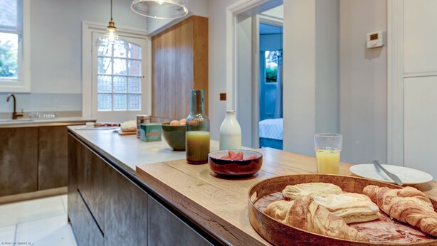 A modern kitchen with a wooden countertop, a large bowl of croissants, a bottle of orange juice, and a glass of orange juice. - Rawlinson Road 10B | Oxford Holiday Let | 2 Beds & Sleeps 4 (Oxford)