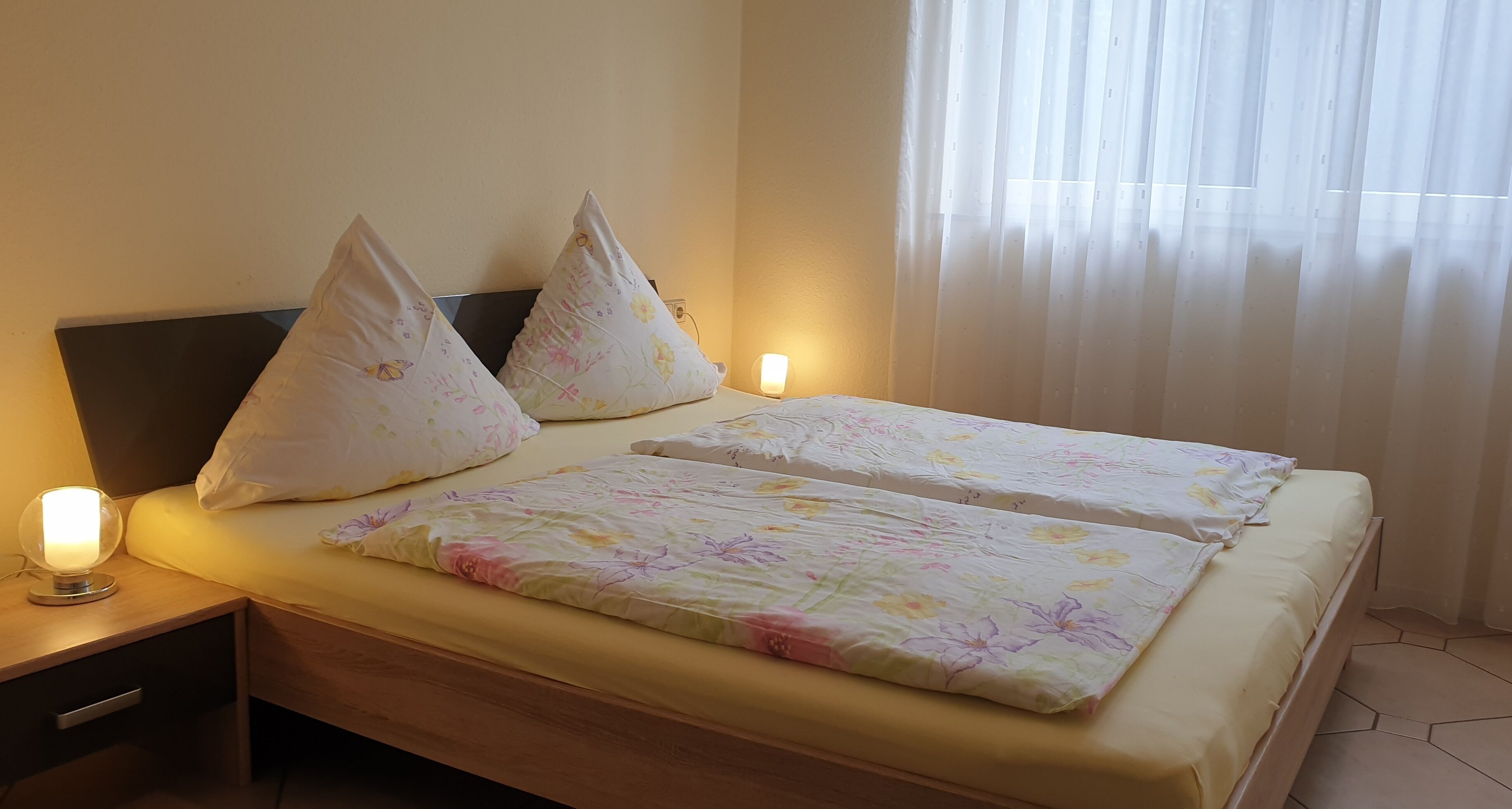 A double bed with floral bedding, two pillows, bedside lamps, and a nightstand with a drawer.
