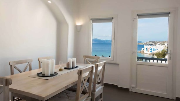 A dining area with a table set for four, two windows offering views of a coastal town, and a door leading outside.