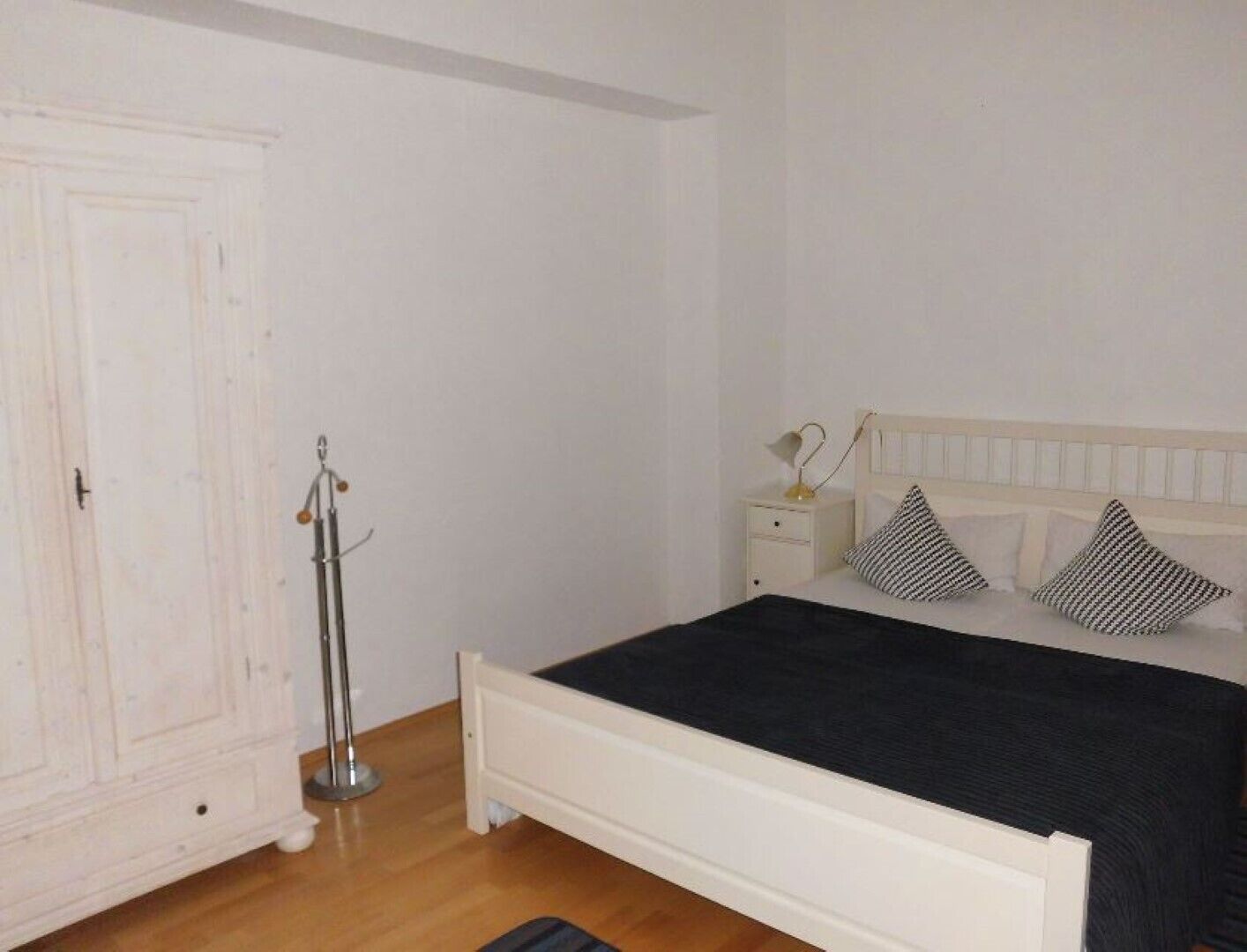 A bedroom with a white bed, a bedside table with a lamp, and a wardrobe.