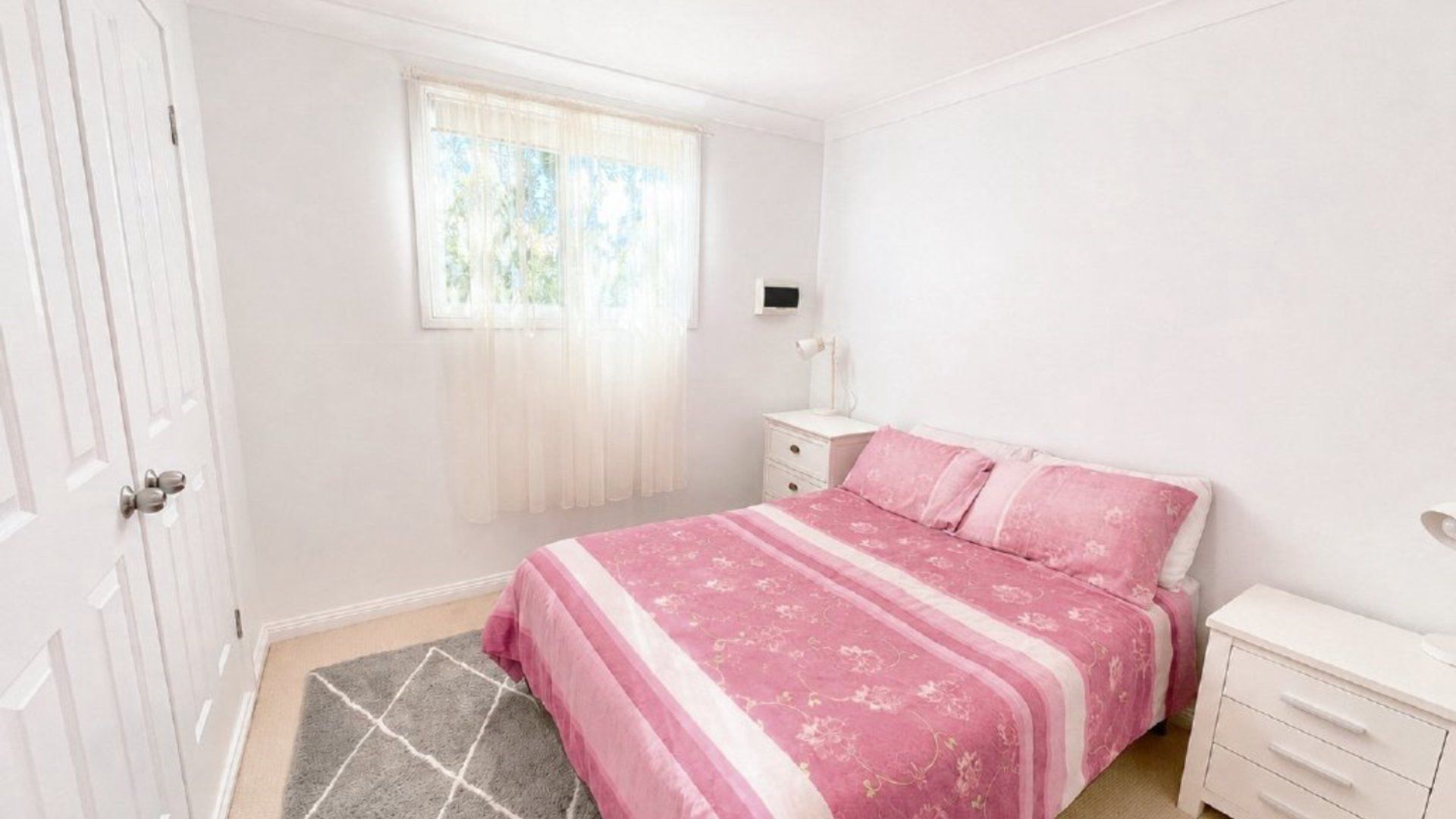 A bedroom with a bed, a nightstand, a desk, a chair, and a window with curtains.