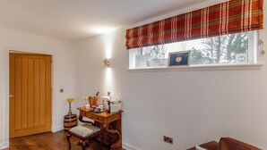 Una habitación con puerta de madera, un escritorio con lámpara, una silla, un ventanal con cortina escocesa y un certificado en la pared.