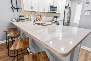 A kitchen with white cabinets, a stainless steel refrigerator, a microwave, and a sink. There are wooden stools and a wooden bench. - Anchorage II 205  Lovely 2 Bedroom Condo With Great Ocean Views and Outdoor Swimming Pool (MYRTLE BEACH)
