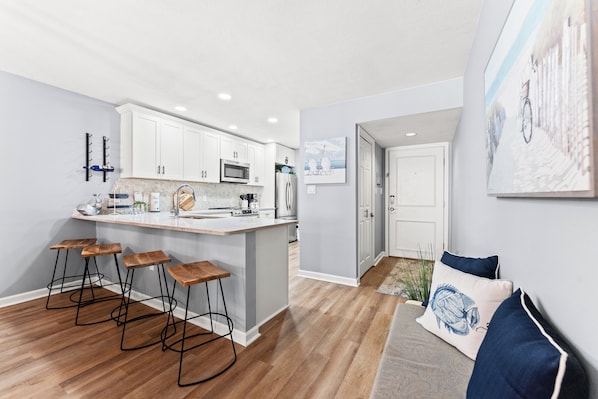 A modern kitchen with white cabinets, a central island, bar stools, and a dining area with a view of the outdoors.