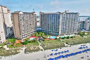 Un hotel con varias construcciones altas, una zona de playa con sombrillas y una piscina.