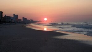 En strand med solnedgang, bygninger i baggrunden og bølger.