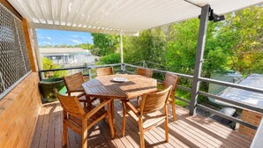 Um deck de madeira com mesa e cadeiras, uma área coberta e vista para o verde e um edifício ao fundo.