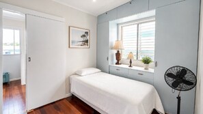 A bedroom with a bed, a window with blinds, a bedside table with a lamp, a fan, and a framed picture on the wall.