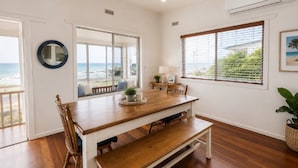 Uma sala de jantar com mesa de madeira, cadeiras e um banco. Há janelas com vista para a praia e o oceano.