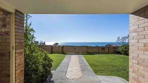 A view from a porch with a wooden fence, a paved pathway, and a grassy yard leading to a body of water.