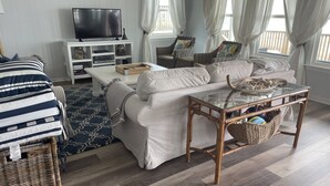 A living room with a sofa, a glass coffee table, a television on a stand, wicker furniture, and a patterned rug. - Beachfront Home! Ready for the summer! (Galveston)