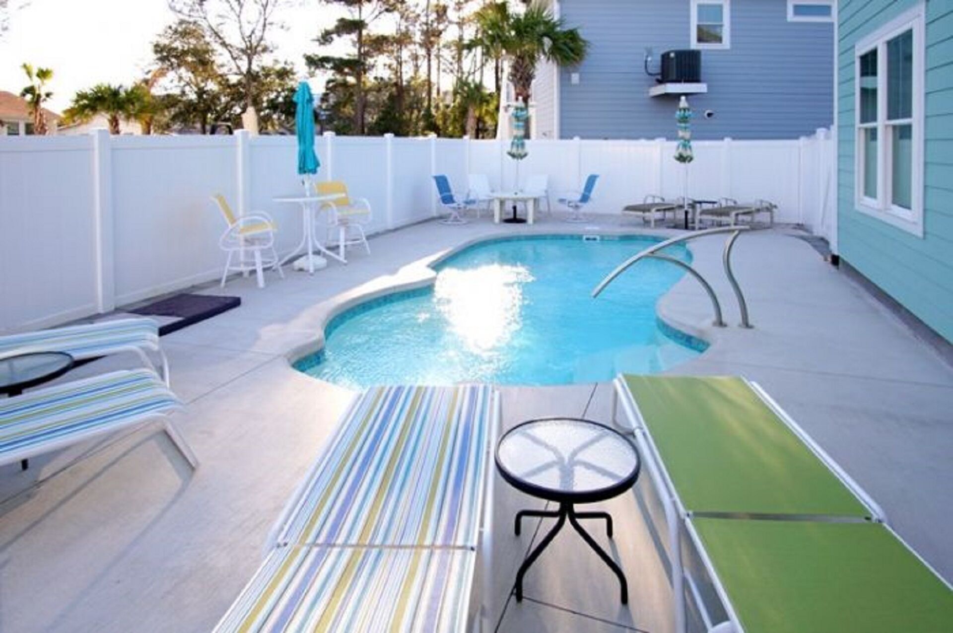 A swimming pool with lounge chairs and a table, surrounded by a white fence and trees.