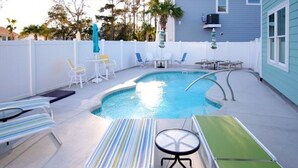 A swimming pool with lounge chairs and a table, surrounded by a white fence and trees.