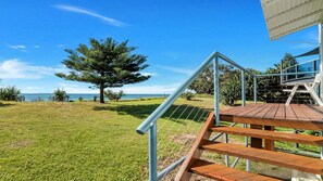 A wooden deck with a metal railing, overlooking a grassy area with a tree and a view of the sea.