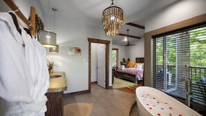 Un baño moderno con bañera, ducha y tocador. Se ve una puerta que conduce a otra habitación y una ventana con persianas.