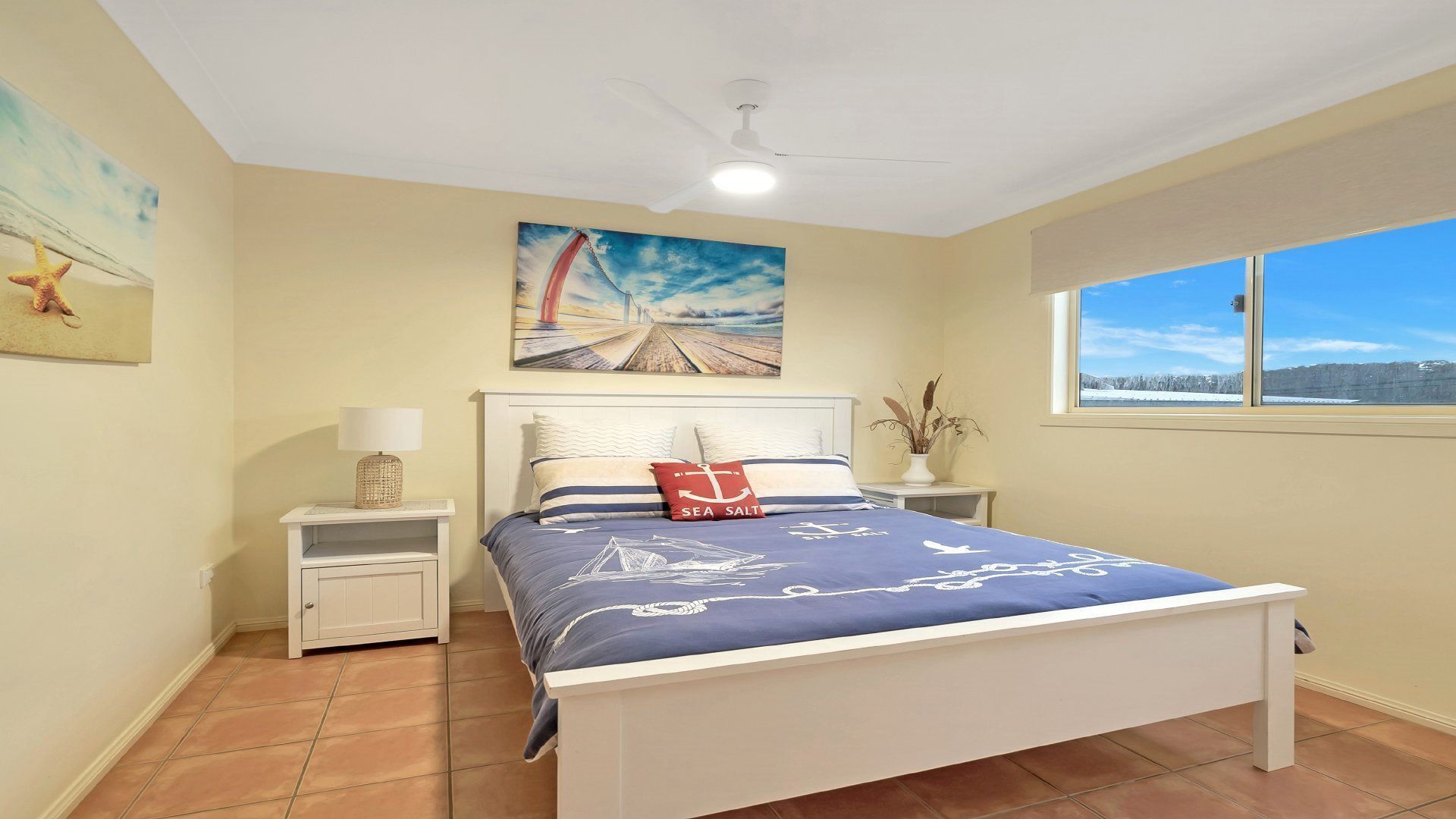 Un dormitorio con cama, mesita de noche, lámpara, un cuadro y ventana con vistas.