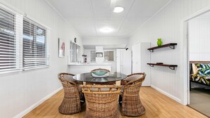 A dining area with a round table, four wicker chairs, white walls, a kitchen in the background, and a door leading to another room. - The Purple Cottage - Burrum Heads - Walk To Beach -2BR -... (BURRUM HEADS)