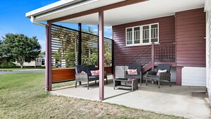 A covered patio with wicker furniture and a staircase leading to a house with a dark exterior. - The Purple Cottage - Burrum Heads - Walk To Beach -2BR -... (BURRUM HEADS)