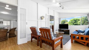 A spacious living area with a TV, wooden furniture, and a view of greenery outside. - The Purple Cottage - Burrum Heads - Walk To Beach -2BR -... (BURRUM HEADS)