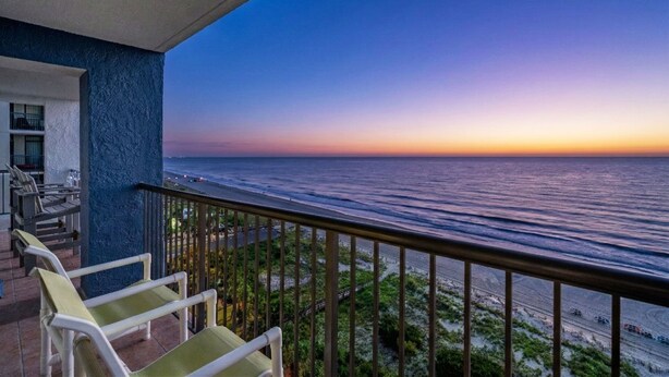 A balcony with chairs overlooking a beach at sunset.