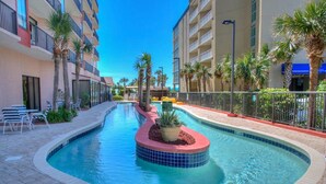 A swimming pool with a tiled hot tub, surrounded by palm trees and a building with balconies.