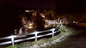 Une vue nocturne d’un quartier résidentiel, avec une clôture blanche, un jardin bien éclairé et une maison dont on aperçoit les fenêtres et la porte.