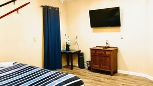 Ein Schlafzimmer mit einem Bett, einem an der Wand montierten Fernseher, einem Holzschrank und einem Schreibtisch mit Pflanze.