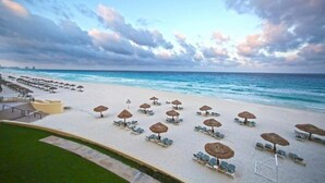 A beach with rows of sun loungers and thatched umbrellas, a clear view of the ocean, and a distant city skyline. - The Royal Sands All Inclusive Resort & Spa (Cancun)