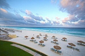 A beach with rows of sun loungers and thatched umbrellas, a clear view of the ocean, and a distant city skyline. - The Royal Sands All Inclusive Resort & Spa (Cancun)