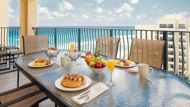 Una mesa de desayuno con waffles, fruta y jugo en un balcón con vista al océano.