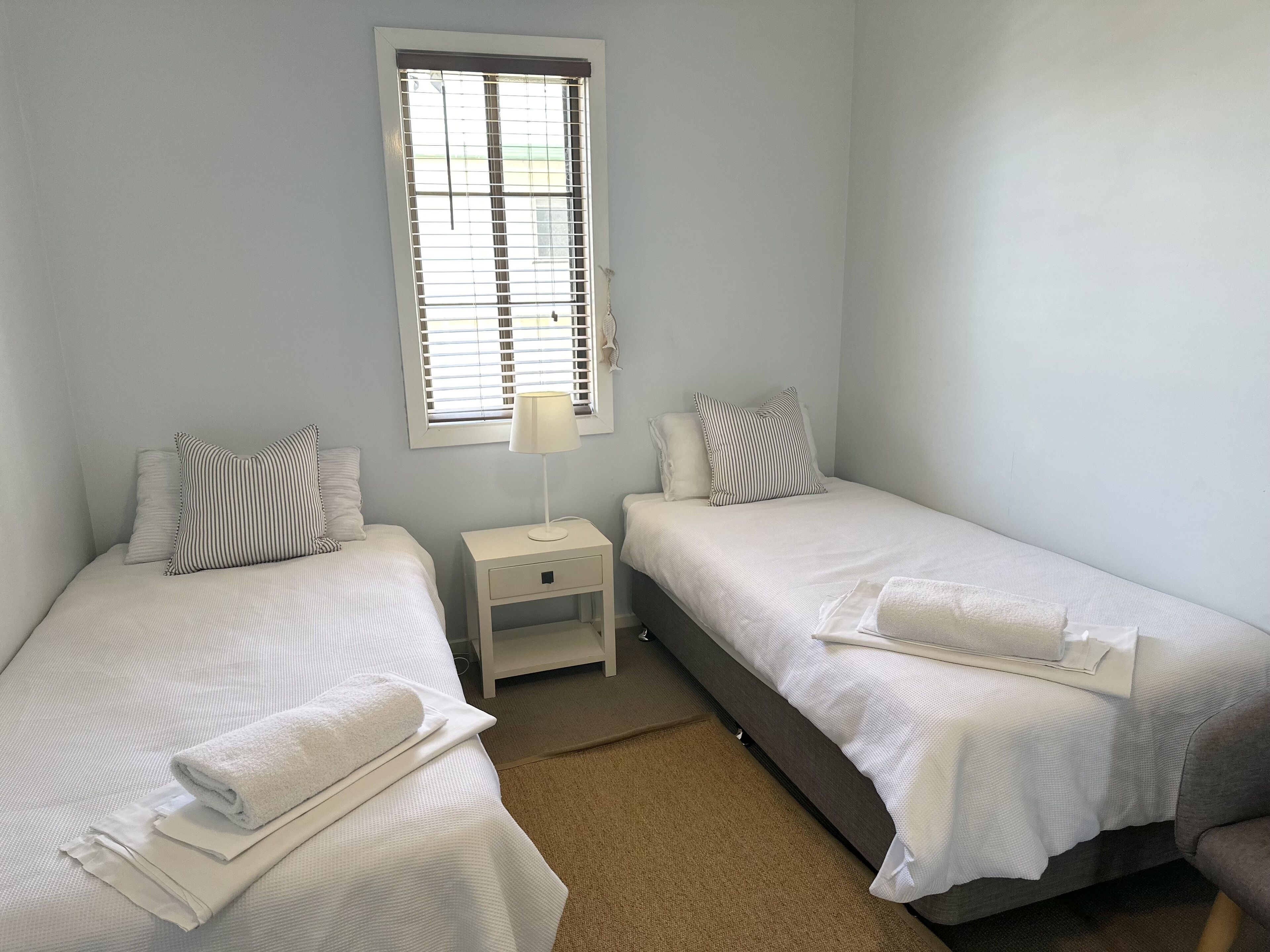 A bedroom with two single beds, a bedside table with a lamp, and a window with blinds.