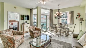A spacious living room with a glass coffee table, floral armchairs, a dining area with a chandelier, and a balcony with a view. - Light and Spacious Corner Unit, Ocean View, Pools | Crescent Keyes - PH 10 (North Myrtle Beach)