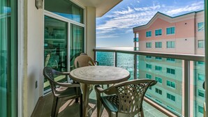 A balcony with a round table and two chairs, overlooking a body of water and a building with multiple windows. - Light and Spacious Corner Unit, Ocean View, Pools | Crescent Keyes - PH 10 (North Myrtle Beach)