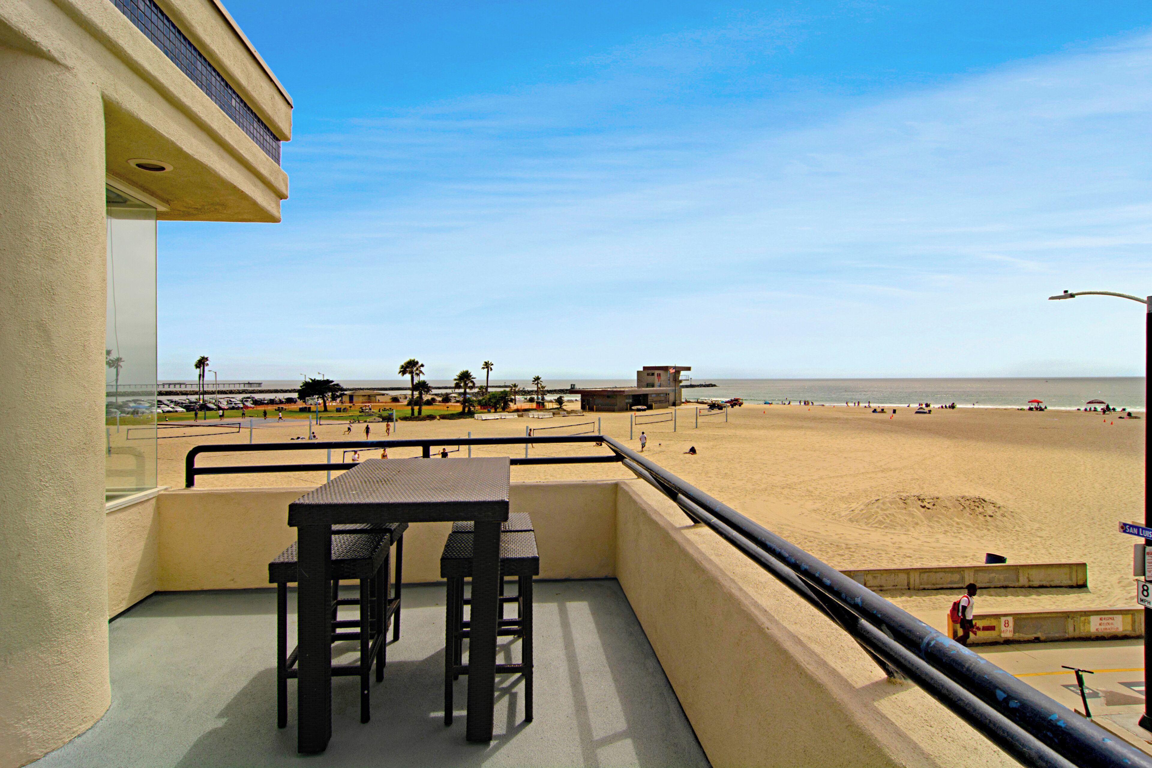 Un balcon avec une table et des chaises donnant sur une plage, où l’on aperçoit une tour de surveillance et des personnes se baignant.