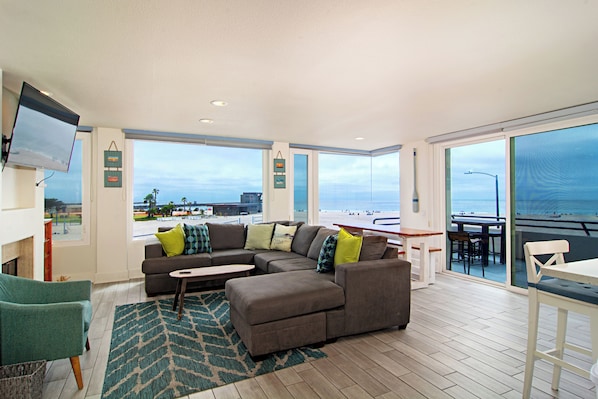 A spacious living area with a large window overlooking a beach, a sofa with multiple cushions, a small table, and a television mounted on the wall.