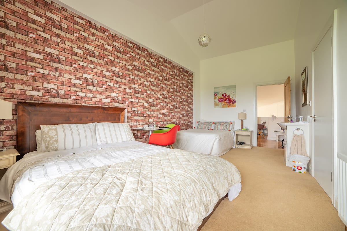 A bedroom with a brick wall, a large bed with a quilted duvet, a red chair, and a small desk with a chair.
