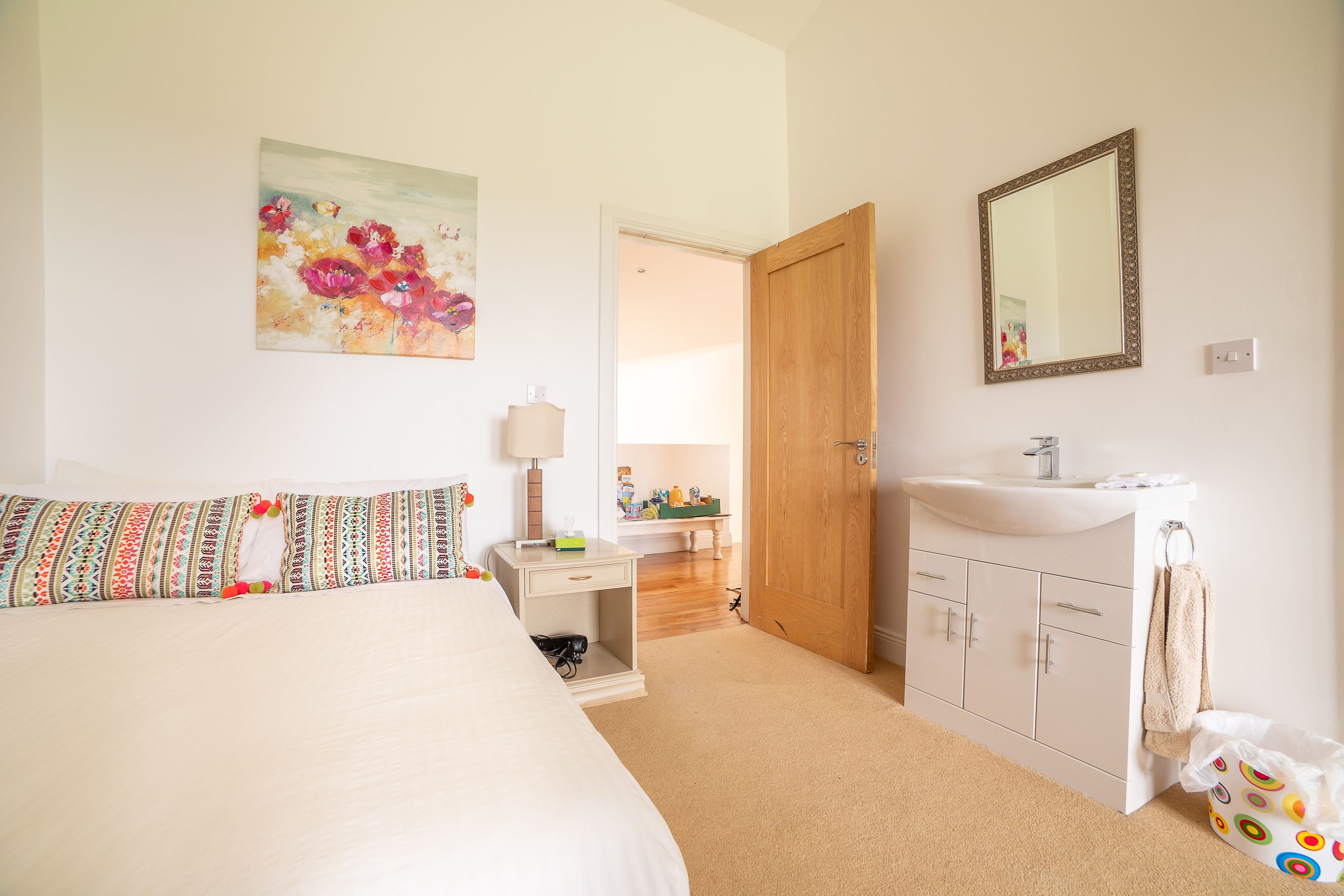 A bedroom with a bed, a bedside table with a lamp, a dresser, and a bathroom with a sink and mirror.