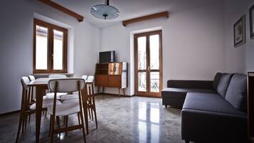 Una sala de estar con comedor, un sofá, un televisor sobre un mueble de madera y ventanas grandes.
