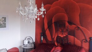 Un dormitorio con cama, lámpara de araña, pared roja y una mesita de noche pequeña.